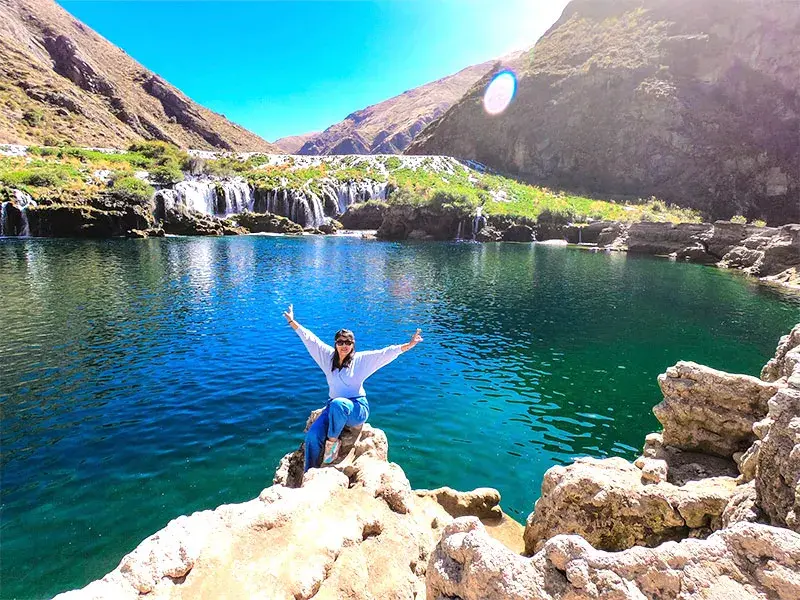 Viajera sentada sobre rocas frente a una gran laguna turquesa y cascadas naturales durante un full day huancaya en la sierra de Lima.